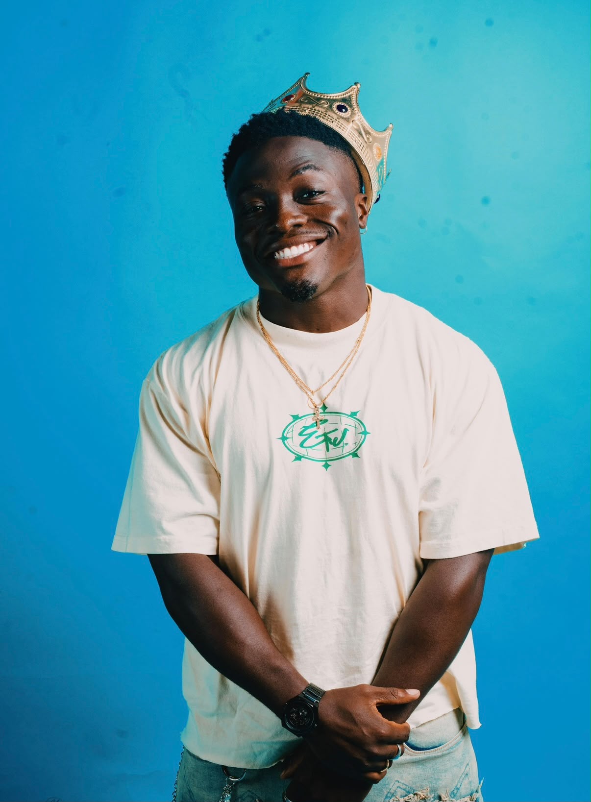 Call Me Manny smiling in a crown against a bright blue backdrop