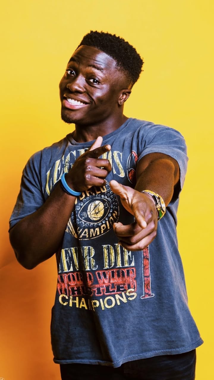 Call Me Manny grinning and pointing toward the camera against a yellow backdrop